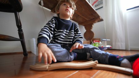 Téléchargez les photos : Un petit garçon assis sur un plancher de bois franc, rassemblant des rails de train en bois. Bâtiment enfant piste rétro - en image libre de droit