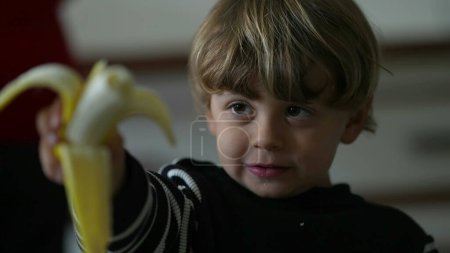 Photo for Portrait of little boy eating banana, close-up face of a caucasian blonde child snacking healthy fruit, candid lifestyle - Royalty Free Image