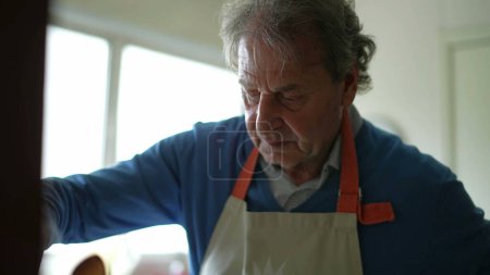 Foto de Primer plano del hombre mayor cocinando alimentos por cocina estufa, anciano jubilado en los años 70 preparando la comida y el uso de delantal - Imagen libre de derechos