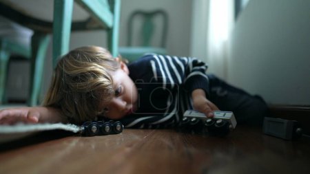 Téléchargez les photos : Enfant jouant par lui-même sur le sol à la maison, un petit garçon engagé avec son propre monde imaginatif tenant jouet de voiture à la main, posé sur le plancher de bois franc - en image libre de droit
