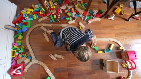 Photo for Small boy playing with toys seen from above perspective, child immersed in play with retro vintage railroad tracks and blocks scattered around - Royalty Free Image