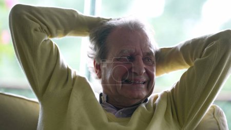 Foto de Feliz hombre mayor escuchando la conversación mientras está sentado en el sofá con las manos apretadas detrás de la cabeza sonriendo - Imagen libre de derechos