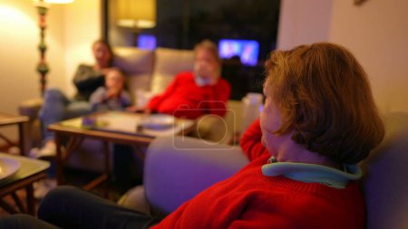 Photo for Senior woman watching television seated on couch, family in background. Boomer generation lady staring at TV screen entertainment media - Royalty Free Image