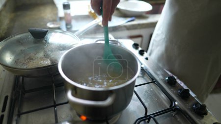 Photo for Close-up hand stirring food in boiling water inside metal pan at kitchen stove, meal preparation - Royalty Free Image