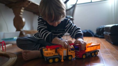 Foto de Pequeño niño jugando con sus juguetes de camiones, golpeando los coches del vehículo juntos. Niño absorto con el juego en casa, niño caucásico macho juega con objetos - Imagen libre de derechos
