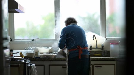 Foto de Candid cocinero senior de pie junto al mostrador de la cocina preparando la comida, hombre mayor genuino preparando la comida vertiendo agua hirviendo en el fregadero, rutina culinaria doméstica diaria - Imagen libre de derechos