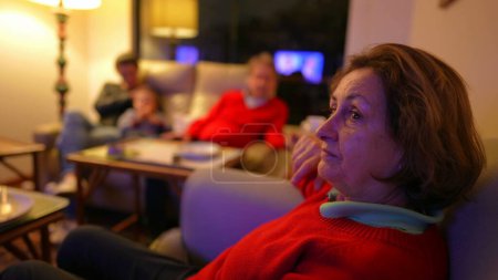 Photo for Senior woman watching television seated on couch, family in background. Boomer generation lady staring at TV screen entertainment media - Royalty Free Image