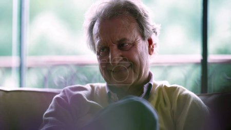 Photo for Thoughtful senior man seated at home couch listening to conversation. Candid elderly man in 70s remaining quiet while he listens to people talk - Royalty Free Image