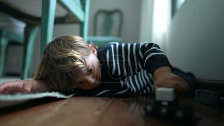 Téléchargez les photos : Enfant jouant par lui-même sur le sol à la maison, un petit garçon engagé avec son propre monde imaginatif tenant jouet de voiture à la main, posé sur le plancher de bois franc - en image libre de droit