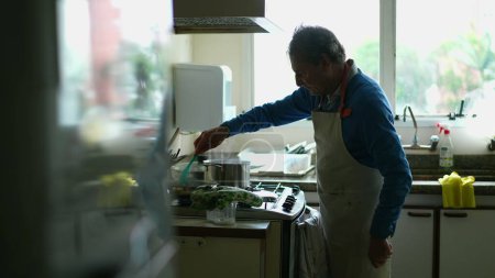Foto de Hombre mayor cándido cocinar alimentos por cocina estufa usando delantal. Una persona anciana caucásica preparando comida para la familia - Imagen libre de derechos