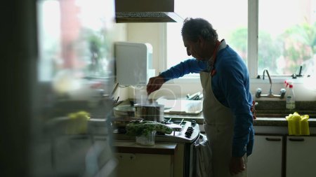 Foto de Hombre mayor cándido cocinar alimentos por cocina estufa usando delantal. Una persona anciana caucásica preparando comida para la familia - Imagen libre de derechos