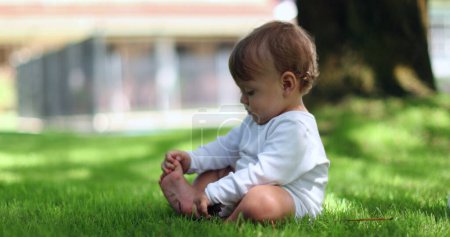 Foto de Lindo bebé sentado afuera jugando con palo y dedos de los pies en el césped - Imagen libre de derechos