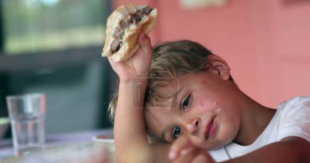 Téléchargez les photos : Mignon garçon tout-petit ennuyé manger hamburger pour le déjeuner. L'enfant mange un hamburger - en image libre de droit