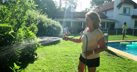 Téléchargez les photos : Senior woman watering plants with water hose outside. Relaxed older lady in 60s taking care of garden - en image libre de droit