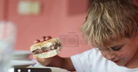 Téléchargez les photos : Jeune garçon tenant hamburger à la chaise de réglage du déjeuner - en image libre de droit
