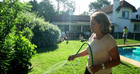 Foto de Older woman watering plants in residential home backyard garden with water hose - Imagen libre de derechos