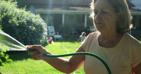 Photo for Older woman gardening and water plants with water hose outside in sunlight - Royalty Free Image