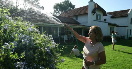 Téléchargez les photos : Older woman watering plants during sunny beautiful day with water hose. Residential house backyard with family relaxing - en image libre de droit