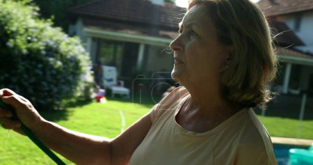 Téléchargez les photos : Woman in 60s watering plants with water hose outdoors with sunlight lens-flare - en image libre de droit