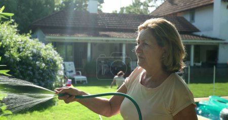 Téléchargez les photos : Older woman gardening and water plants with water hose outside in sunlight - en image libre de droit