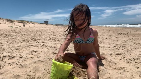 Téléchargez les photos : Enfant fille à la plage jouant avec le sable remplissage seau avec du sable - en image libre de droit