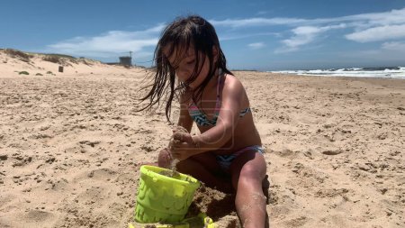 Téléchargez les photos : Enfant fille à la plage jouant avec le sable remplissage seau avec du sable - en image libre de droit