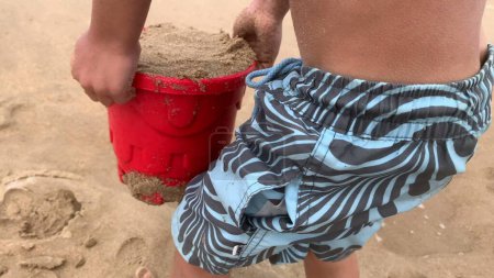 Téléchargez les photos : Enfant garçon portant des seaux remplis de sable à la plage, bâtiment château de sable - en image libre de droit