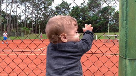 Photo for Cute baby holding into tennis court fence looking at siblings play game - Royalty Free Image
