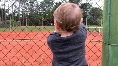 Photo for Cute baby holding into tennis court fence looking at siblings play game - Royalty Free Image