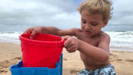 Foto de Lindo niño jugando en la playa con cubos y agua - Imagen libre de derechos