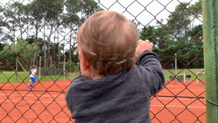 Photo for Baby infant holding into tennis fence watching game - Royalty Free Image