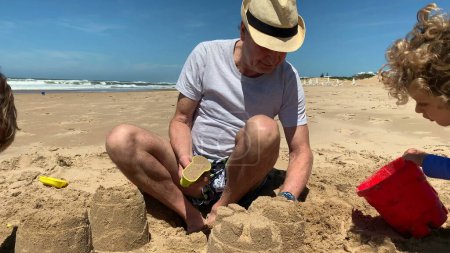 Téléchargez les photos : Grand-père à la plage bâtiment château de sable avec petits-enfants - en image libre de droit