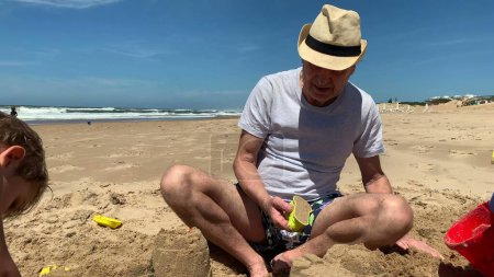 Téléchargez les photos : Grand-père à la plage bâtiment château de sable avec petits-enfants - en image libre de droit