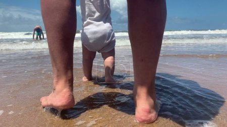 Téléchargez les photos : Bébé bébé mignon pieds nus à la plage, vague s'écrasant à terre. Bébé sentir l'eau de mer - en image libre de droit