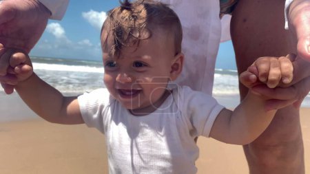 Téléchargez les photos : Bébé mignon à la plage. Parent soutenant le nourrisson à terre, aidant à explorer - en image libre de droit