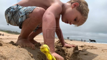 Foto de Niño jugando en la playa. Niño usando la pala para hacer un agujero - Imagen libre de derechos