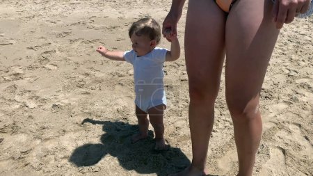 Téléchargez les photos : Bébé enfant apprenant à marcher avec l'aide de la mère se tenant la main à la plage - en image libre de droit
