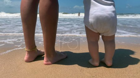 Téléchargez les photos : Bébé à la plage tenant maman par la main. Enfant tout-petit premiers pas au bord de la mer - en image libre de droit