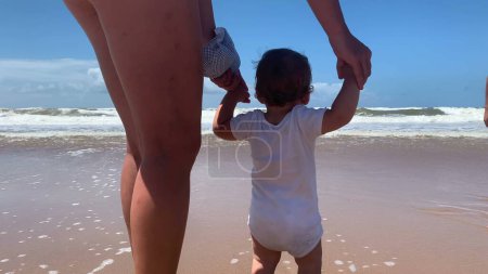 Photo for Baby walking at beach shore for first time, mother helping and holding hands with toddler infant at shore - Royalty Free Image
