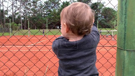 Téléchargez les photos : Bébé mignon tenant dans la clôture du court de tennis en regardant les frères et sœurs jouer au jeu - en image libre de droit
