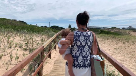 Téléchargez les photos : Jeune mère marchant vers la plage tenant bébé bébé sur le chemin - en image libre de droit