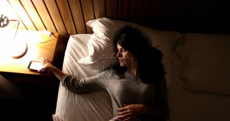 Téléchargez les photos : Woman lying in bed turns off smartphone and bedside nightstand lamp - en image libre de droit