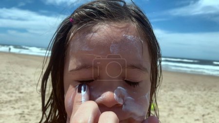 Téléchargez les photos : Parent appliquant de la crème solaire sur le visage de sa fille. Mère frottant et répandant la crème solaire à l'enfant - en image libre de droit