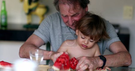 Photo for Baby grandson boy on grandfather lap eating watermelon. Infant refusing food, not wanting to eat - Royalty Free Image