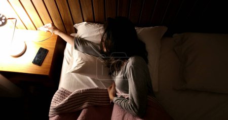 Téléchargez les photos : Young woman lies in bed going to sleep, turning night-stand light lamp off - en image libre de droit