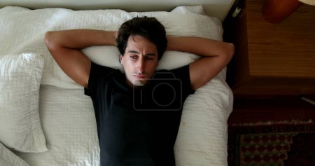 Téléchargez les photos : Pensive young man lying in bed resting. Thoughtful person lies in bed in thought - en image libre de droit