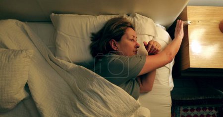 Foto de Thoughtful older woman switching lamp light OFF, person going to sleep - Imagen libre de derechos