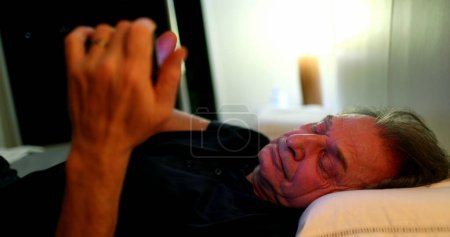 Photo for Older man looking at cellphone screen at night laying down in bed - Royalty Free Image