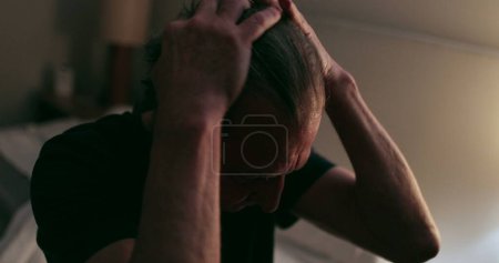 Photo for Old man sitting by the side of the bed at night rubbing face with hands - Royalty Free Image