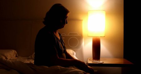 Téléchargez les photos : Pensive older woman sits by the side of the bed preparing to go to sleep - en image libre de droit